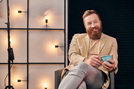 fröhlicher gutaussehender Mann mit rotem Bart in eleganter Kleidung sitzend und glücklich lächelnd beim Interview