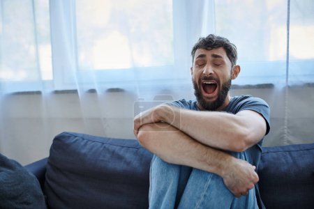 hombre traumatizado deprimido en camiseta casual sentado en el sofá y gritando, conciencia de salud mental