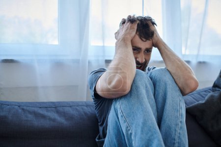 depressiver traumatisierter Mann in lässigem T-Shirt auf Sofa sitzend und schreiend, Bewusstsein für psychische Gesundheit