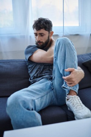 Foto de Hombre ansioso traumatizado en ropa casual sentado en el sofá durante la ruptura, conciencia de salud mental - Imagen libre de derechos
