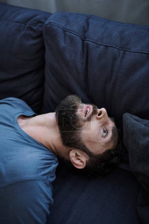 verzweifelter depressiver Mann in lässigem T-Shirt während depressiver Episode auf Sofa liegend, psychische Gesundheit
