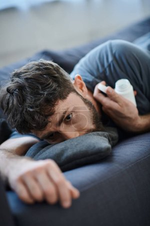 Foto de Traumatizado hombre sufriente con barba acostado en el sofá con pastillas en la mano, conciencia de la salud mental - Imagen libre de derechos