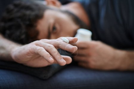 Photo for Depressed anxious man in casual attire lying on sofa with pills in hand, mental health awareness - Royalty Free Image