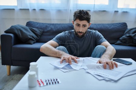 Foto de Hombre deprimido enfermo con barba sentado en la mesa con papeles y pastillas en él durante el episodio depresivo - Imagen libre de derechos