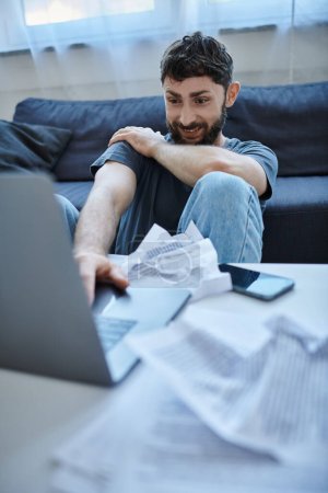 Foto de Hombre deprimido con sonrisa loca en la ropa de casa mirando a su computadora portátil durante la crisis, la salud mental - Imagen libre de derechos