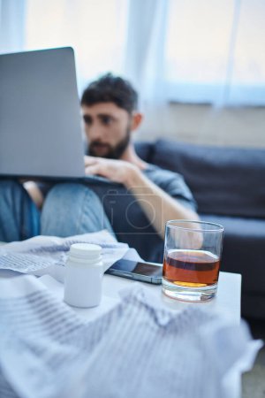 hombre traumatizado deprimido con barba trabajando en el ordenador portátil con un vaso de bebida alcohólica en la mesa