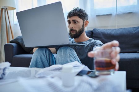 hombre traumatizado deprimido con barba trabajando en el ordenador portátil con un vaso de bebida alcohólica en la mesa