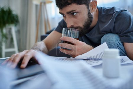 Verzweifelter traumatisierter Mann mit Bart arbeitet am Laptop und trinkt Alkohol auf dem Tisch