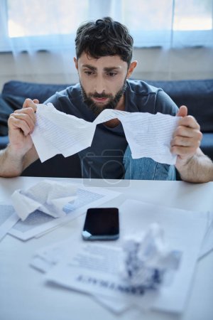 Photo for Frustrated man with beard sitting at table with papers and phone on it during mental breakdown - Royalty Free Image