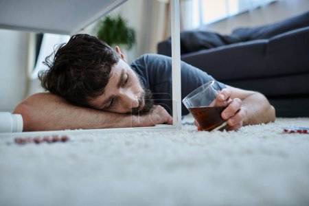 Depressiver Mann in Freizeitkleidung trinkt Alkohol und schaut bei psychischem Zusammenbruch auf Tabletten
