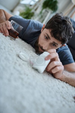 Photo for Frustrated ill man in casual attire drinking alcohol and holding pills during mental breakdown - Royalty Free Image