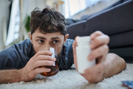 Frustrierter Kranker in Freizeitkleidung trinkt Alkohol und hält Tabletten während des Nervenzusammenbruchs in der Hand