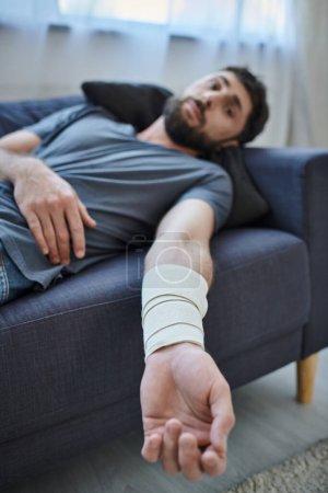 Traumatisierter Mann mit Verband am Arm nach Selbstmordversuch auf Sofa liegend, Bewusstsein für psychische Gesundheit