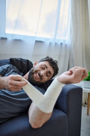 Foto de Hombre enfermo con sonrisa y vendaje en el brazo después de intentar suicidarse en el sofá, conciencia de salud mental - Imagen libre de derechos