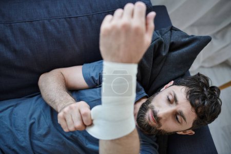 Foto de Hombre traumatizado con vendaje en el brazo después de intentar suicidarse acostado en el sofá, conciencia de salud mental - Imagen libre de derechos