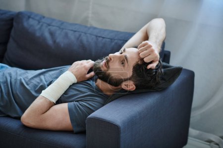 Verzweifelter Mann mit Verband am Arm nach Selbstmordversuch auf Sofa liegend, Bewusstsein für psychische Gesundheit