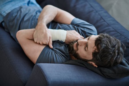 Foto de Hombre desesperado con vendaje en el brazo después de intentar suicidarse acostado en el sofá, conciencia de salud mental - Imagen libre de derechos