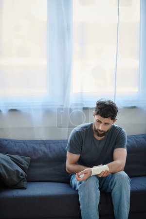 Depressiver Mann mit Verband am Arm nach Selbstmordversuch auf Sofa sitzend, Bewusstsein für psychische Gesundheit