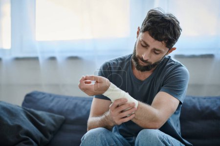 Foto de Hombre deprimido con vendaje en el brazo después de intentar suicidarse sentado en el sofá, conciencia de salud mental - Imagen libre de derechos