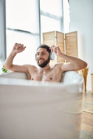 cheerful handsome man with beard and headphones sitting and relaxing in his bathtub, mental health