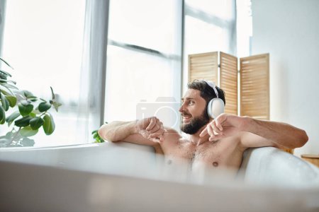 fröhlicher attraktiver Mann mit Bart und Kopfhörer, der in seiner Badewanne sitzt und entspannt, psychische Gesundheit