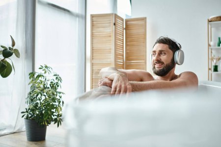 fröhlicher attraktiver Mann mit Bart und Kopfhörer, der in seiner Badewanne sitzt und entspannt, psychische Gesundheit