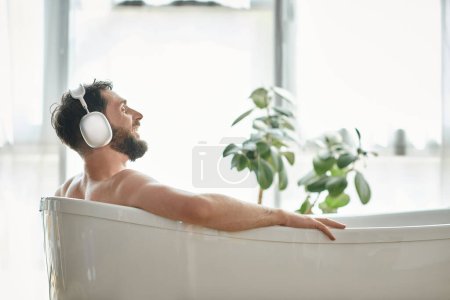 homme joliment beau avec barbe et écouteurs assis et relaxant dans sa baignoire, santé mentale