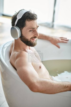 alegre hombre guapo con barba y auriculares sentado y relajante en su bañera, salud mental