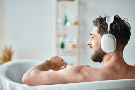 fröhlicher gutaussehender Mann mit Bart und Kopfhörer, der in seiner Badewanne sitzt und entspannt, psychische Gesundheit