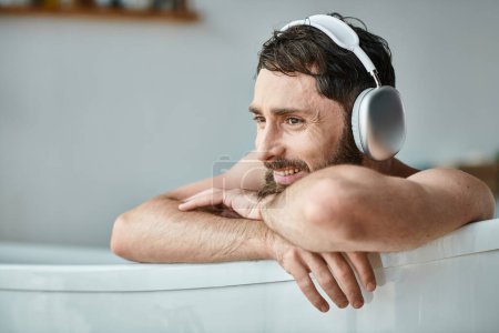 fröhlicher gutaussehender Mann mit Bart und Kopfhörer, der in seiner Badewanne sitzt und entspannt, psychische Gesundheit