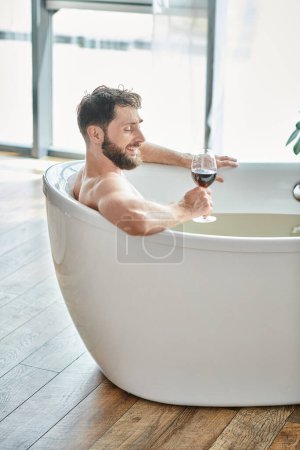 joyous handsome man with beard relaxing in bathtub with glass of red wine, mental health awareness