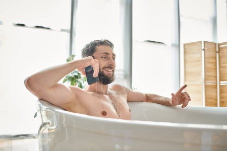 joyous good looking man with beard talking by phone while relaxing in bathtub, mental health