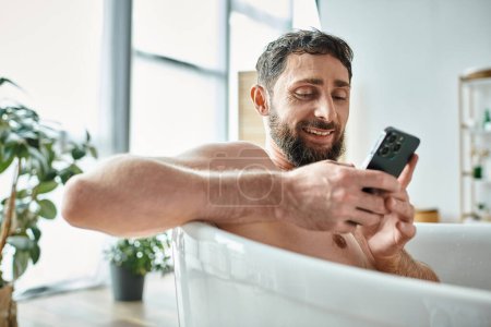 merry handsome man with beard looking at his smartphone while in bathtub, mental health awareness