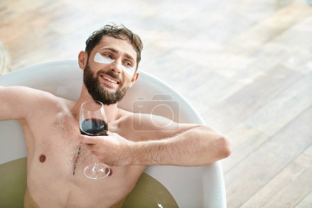 joyful attractive man with beard and eye patches relaxing in bathtub with glass of red wine