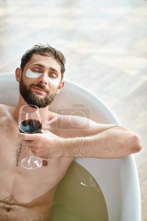 fröhlicher attraktiver Mann mit Bart und Augenklappen entspannt in der Badewanne bei einem Glas Rotwein