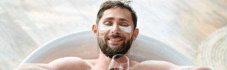 lustiger attraktiver Mann mit Bart und Augenklappen entspannt in der Badewanne bei einem Glas Rotwein, Banner