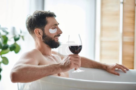 fröhlicher gutaussehender Mann mit Bart und Augenklappen entspannt in der Badewanne bei einem Glas Rotwein