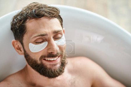 cheerful handsome man with beard relaxing in his bathtub with patches on eyes, mental health