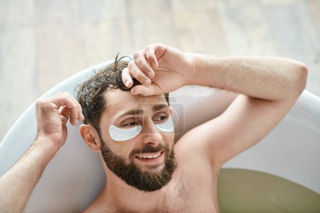 cheerful handsome man with beard relaxing in his bathtub with patches on eyes, mental health