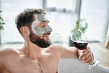 cheerful attractive man with beard and face mask relaxing in bathtub with glass of red wine
