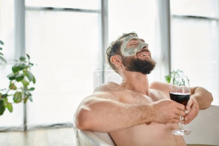 fröhlicher attraktiver Mann mit Bart und Gesichtsmaske entspannt in der Badewanne bei einem Glas Rotwein