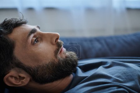 Téléchargez les photos : Homme anxieux déprimé avec barbe en tenue décontractée couché sur le canapé pendant la dépression mentale, la sensibilisation - en image libre de droit