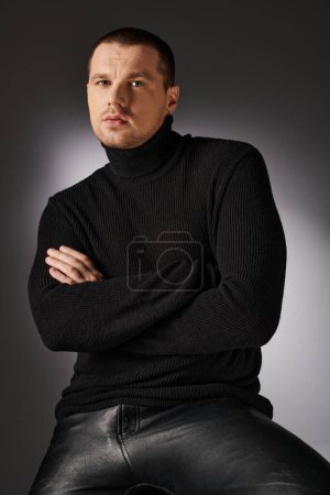 pensive charismatic trendy man in black sweater looking at camera on grey backdrop with lighting