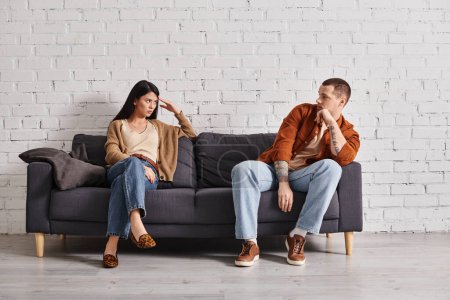 offended interracial couple looking at each other on couch in living room, relationship difficulties