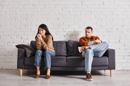 Téléchargez les photos : Jeune homme regardant offensé asiatique femme assis sur canapé dans le salon, relation trouble - en image libre de droit