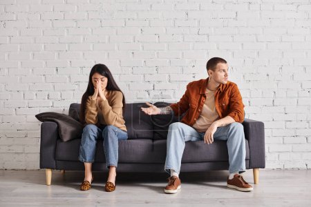 desanimado hombre señalando ofendido asiático esposa en sofá en sala de estar, relación dificultades