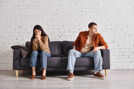 young interracial offended couple sitting on couch in living room, heartache misunderstanding