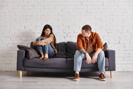 Frustriertes gemischtrassiges Paar schaut einander auf Couch im Wohnzimmer an, Scheidungskonzept