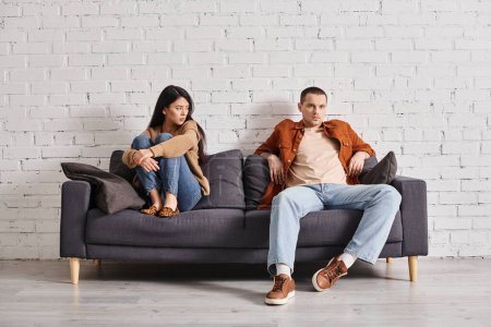 frustrated asian woman sitting near self-assured husband on couch in living room, misunderstanding
