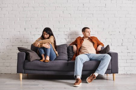 beleidigte asiatische Frau schaut selbstbewussten Ehemann auf Couch im Wohnzimmer an, Missverständnis